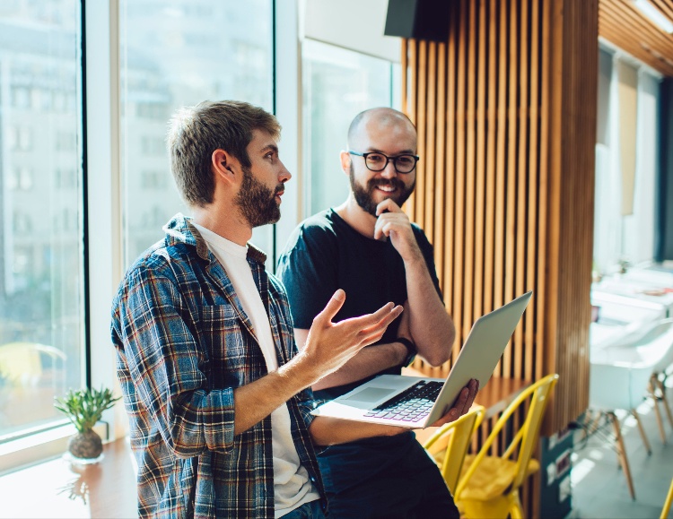 Two men in a modern office engage in conversation by a large window. One, likely an IT partner, holds a laptop while gesturing, and the other listens with a smile. Yellow chairs and a potted plant are visible in the background.