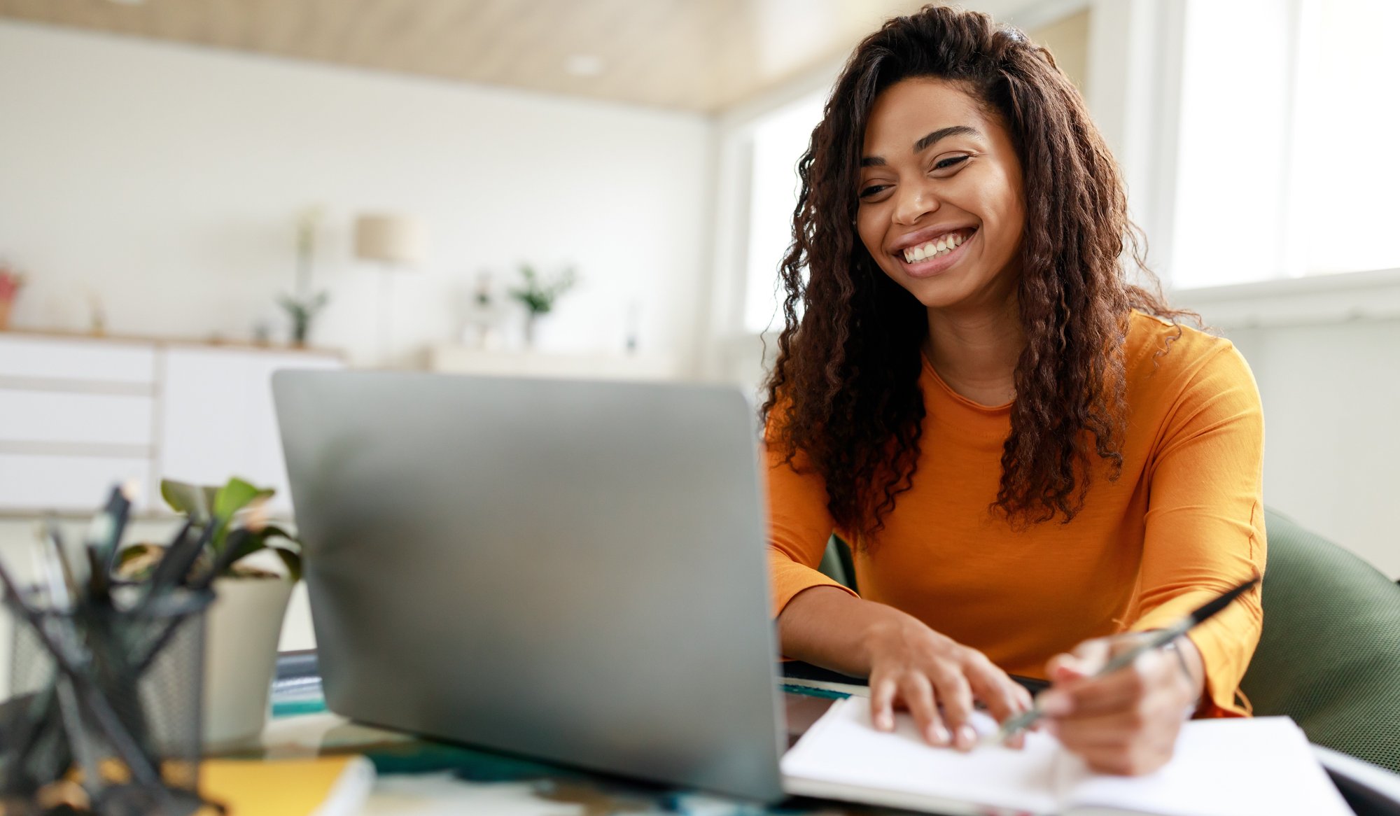 A woman wearing a yellow sweater is smiling while working on a laptop at a desk. She is holding a pen and writing in a notebook. The background shows a bright, modern room with large windows.