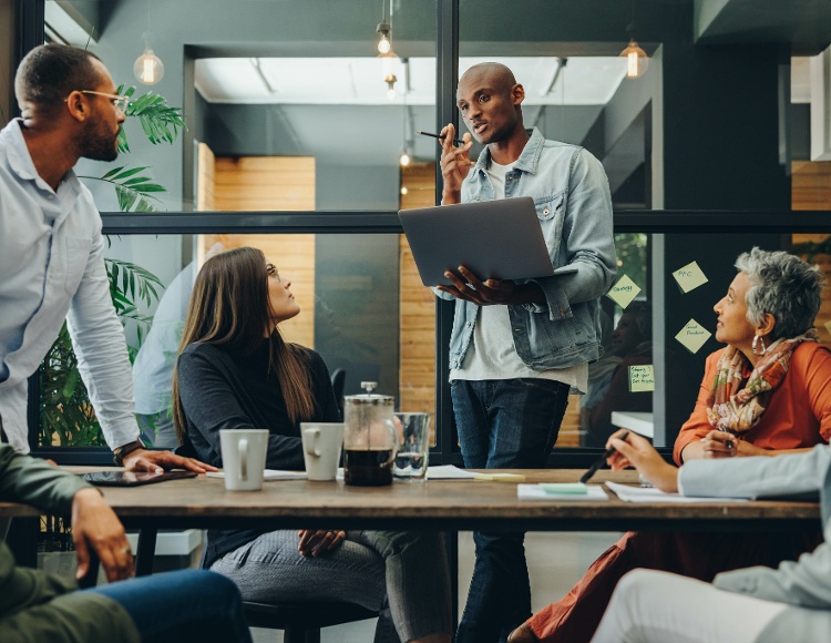 A diverse group of people in a modern office setting is engaged in a meeting. One person, representing their technology partner, stands holding a laptop, speaking to others who are seated around a table with coffee cups and notes. The atmosphere is collaborative and focused.