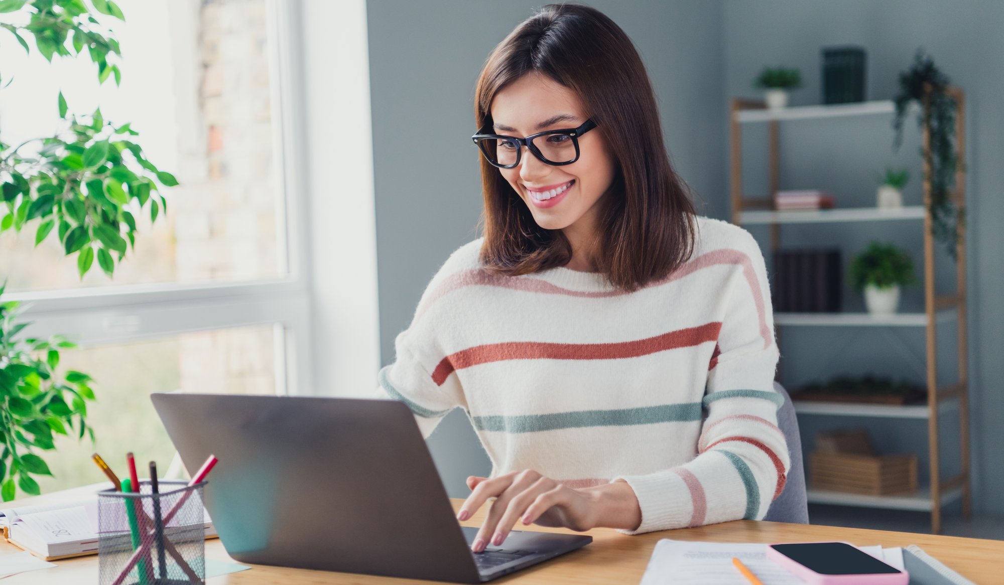 A woman wearing glasses and a striped sweater is smiling while using a laptop at a desk. The desk has notebooks, a pen holder, and a smartphone. A plant and a bookshelf are in the background.