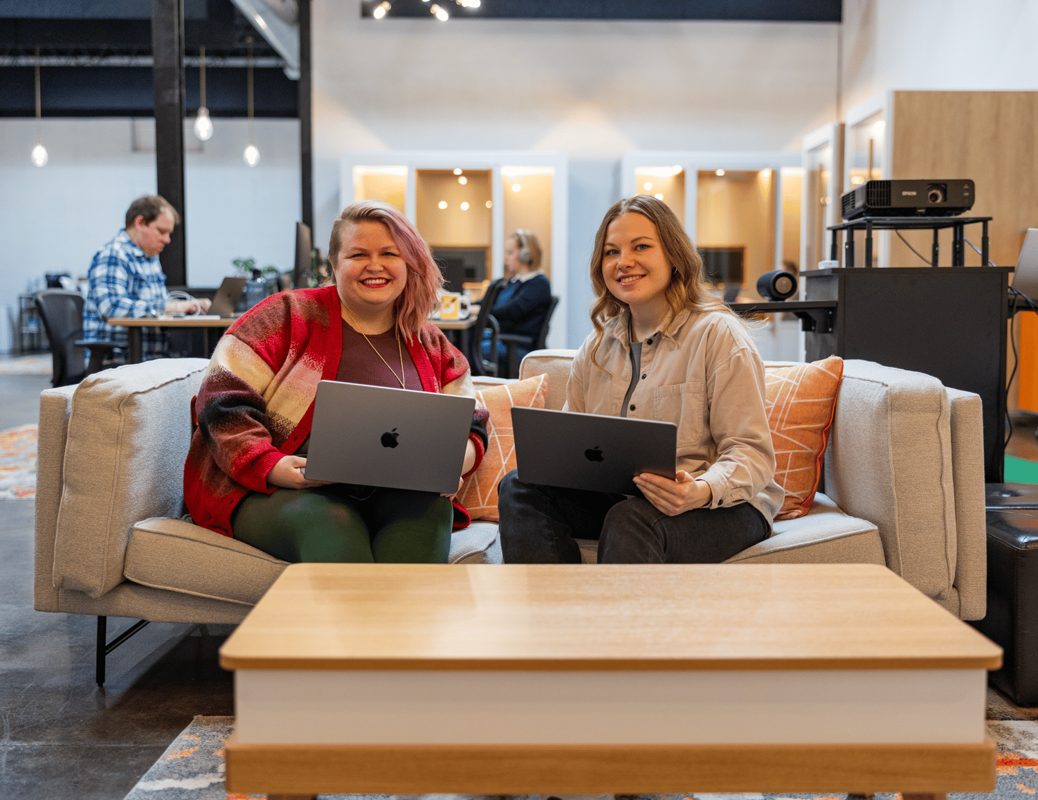 Two people sit on a sofa in an office setting, each holding a laptop and discussing custom software solutions. Both are smiling, with a wooden table before them. In the background, a person works at a desk, surrounded by modern decor.