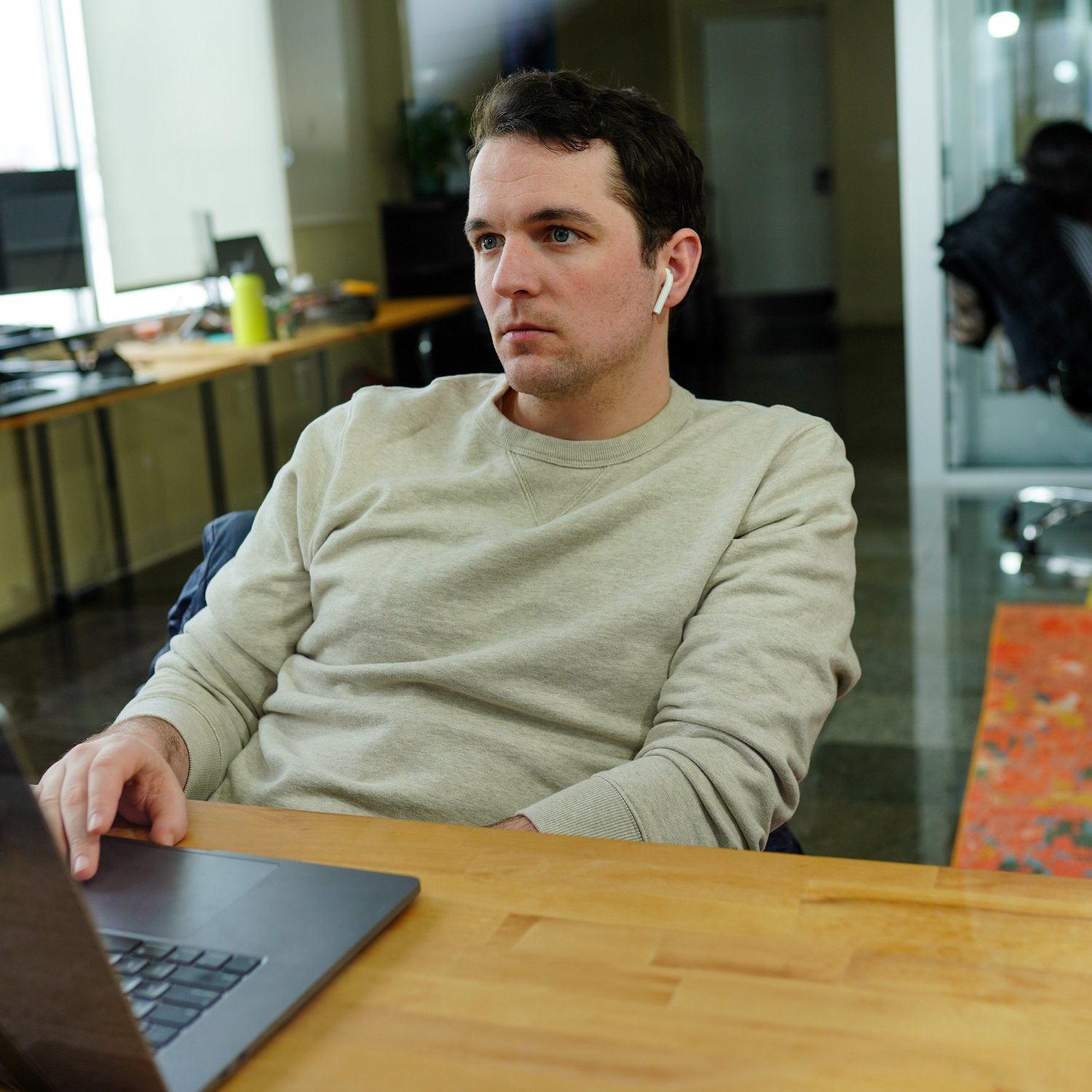 A person sitting at a desk, wearing a beige sweater and wireless earbuds, looking off to the side in an office setting, appears deeply immersed in their software development tasks.