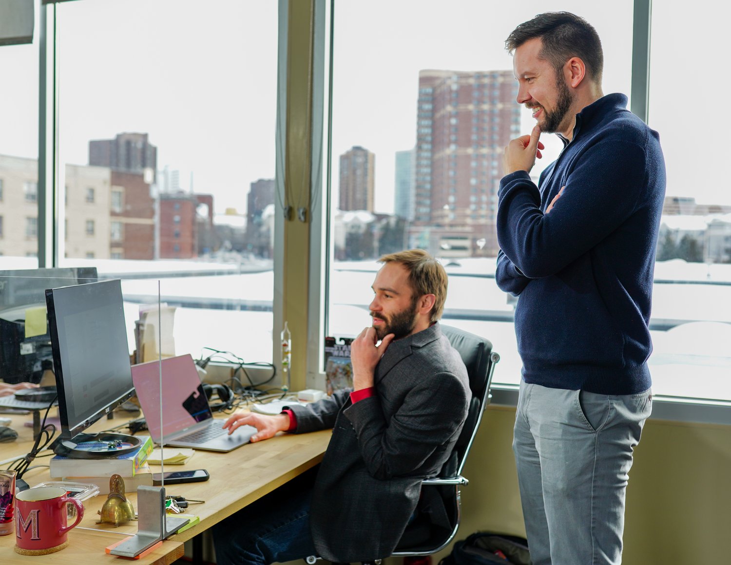 Two men are in an office by a window, looking at a computer screen displaying custom software solutions. One is sitting with his hand on his chin in thought, while the other stands beside him, mirroring the pose. Papers, mugs, and office supplies are scattered on the desk.