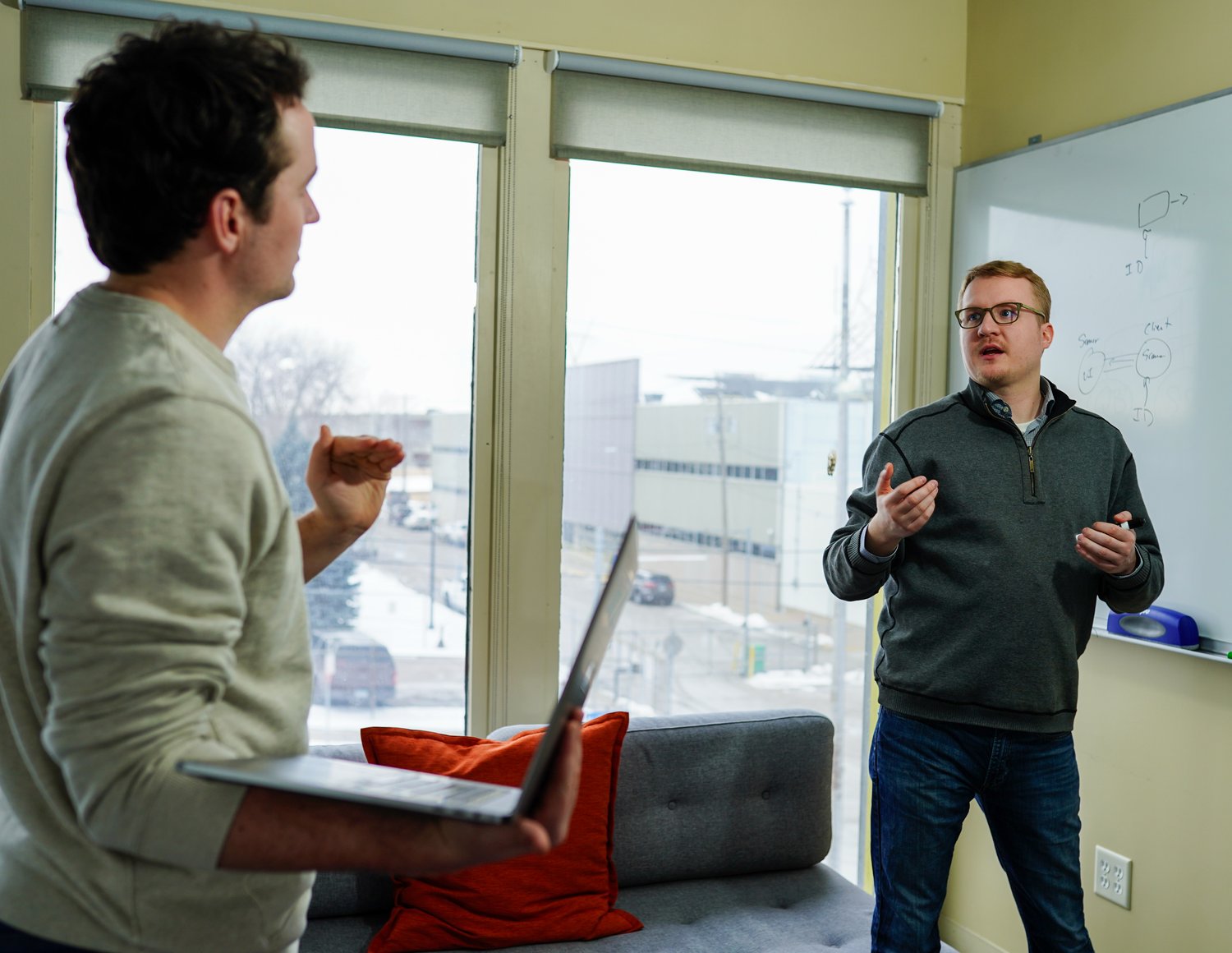 Two people are having a discussion in an office. One is holding a laptop, and the other is standing near a whiteboard with diagrams outlining custom software development solutions. They appear engaged in conversation. The room has large windows with a city view.