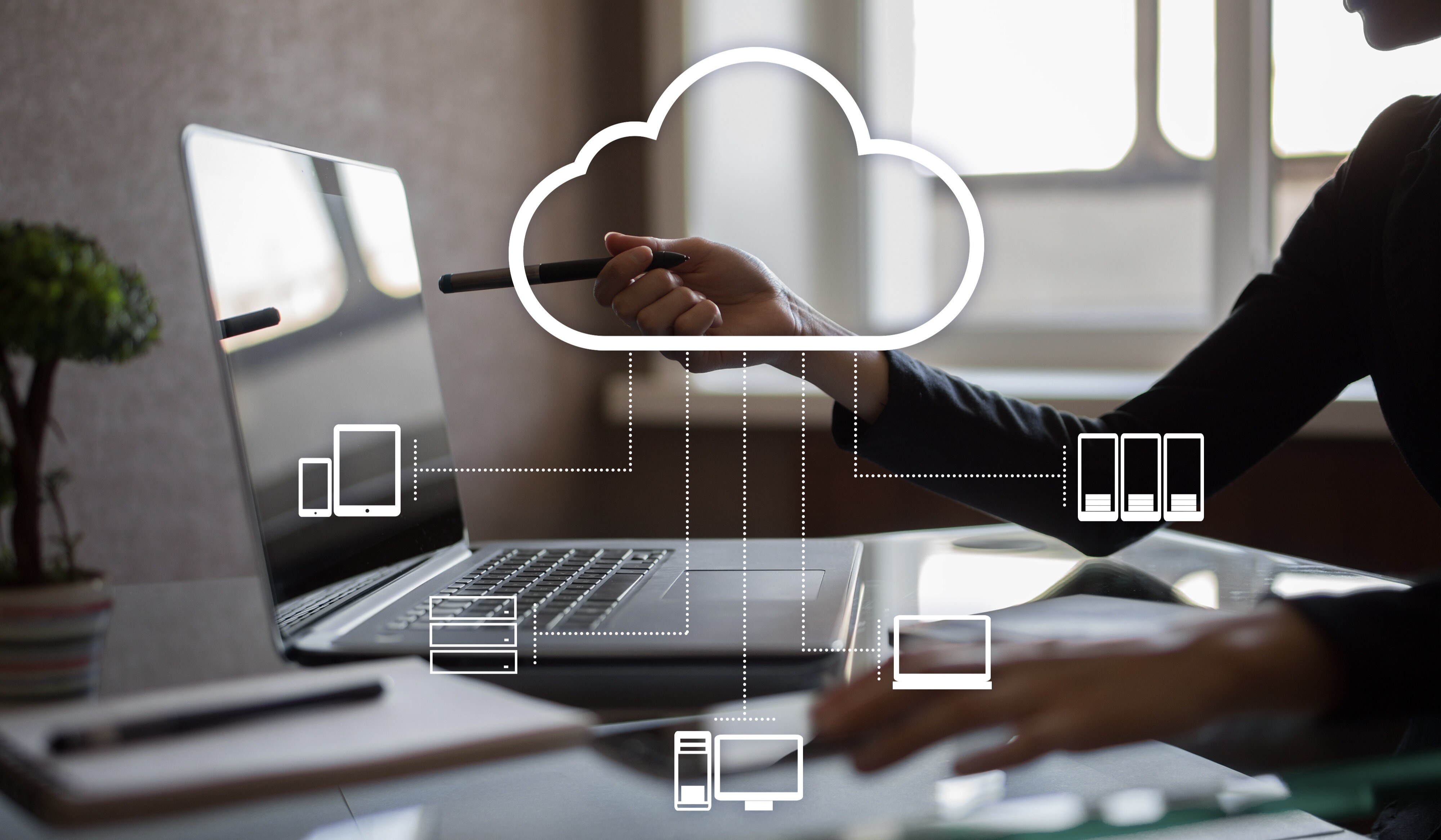 A person points to a laptop screen with a stylus, illustrating modernizing cloud computing. A cloud icon connects to device icons, symbolizing data synchronization in the app. The setting is a modern office bathed in natural light.