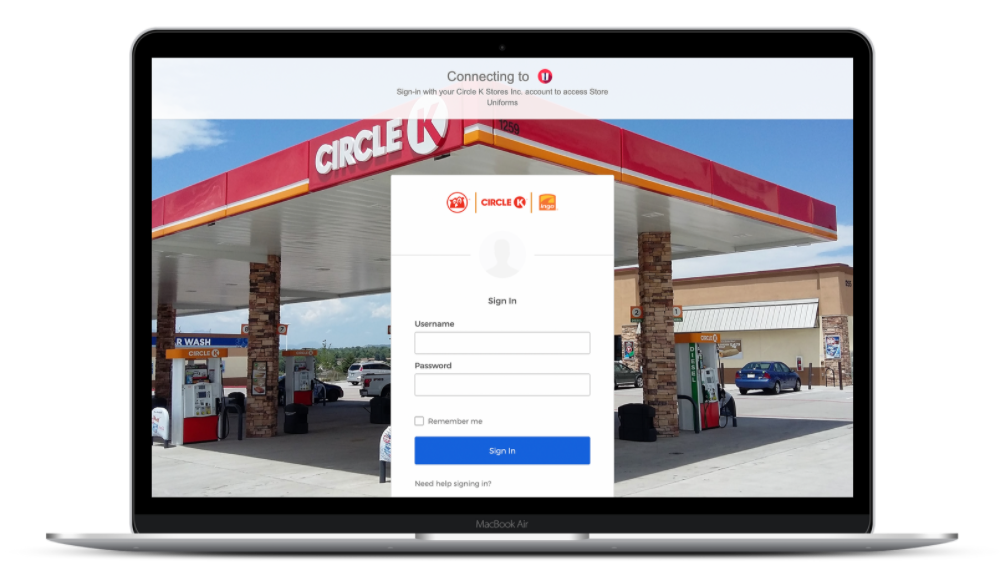 A laptop screen displays a web page for signing into Circle K’s portal, highlighting its robust IT infrastructure with secure username and password fields. In the background, a Circle K gas station is visible, featuring fuel pumps and a car wash area under a red and white canopy.