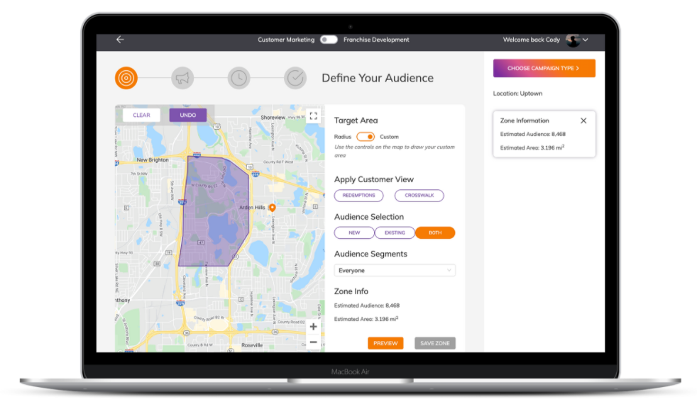 A laptop displaying a marketing tool interface with a map. The map shows a highlighted target area, and options to define audience segments, apply customer view, and choose campaign types. User details are visible at the top.