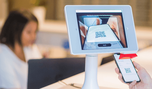 A person uses a smartphone to scan a QR code displayed on a tablet screen. The tablet is mounted on a stand, and a blurred individual works on a laptop in the background, embodying a digital transformation strategy in this modern, tech-oriented environment.