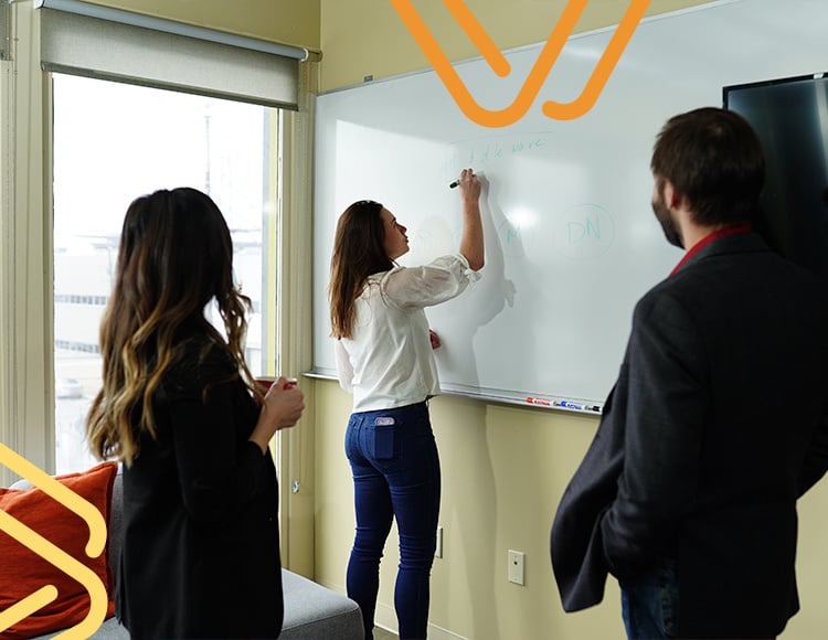 Three people are in a meeting room. One person is writing on a whiteboard while the other two, collaborating as IT partners, watch. A window and a television are in the background. The room is well-lit.