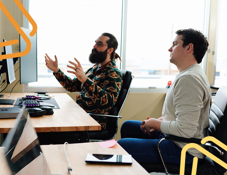 Two men seated at a wooden desk with computer monitors. One gestures while talking, the other listens attentively. In this office setting illuminated by natural light streaming through large windows, they collaborate as technology partners exploring innovative solutions.