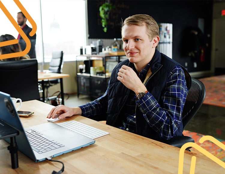 A person wearing a blue plaid shirt and black vest sits at a desk, collaborating with their technology partner while looking at a laptop. The modern workspace, adorned with plants and a large window, creates an inspiring atmosphere. A coffee cup rests beside them.